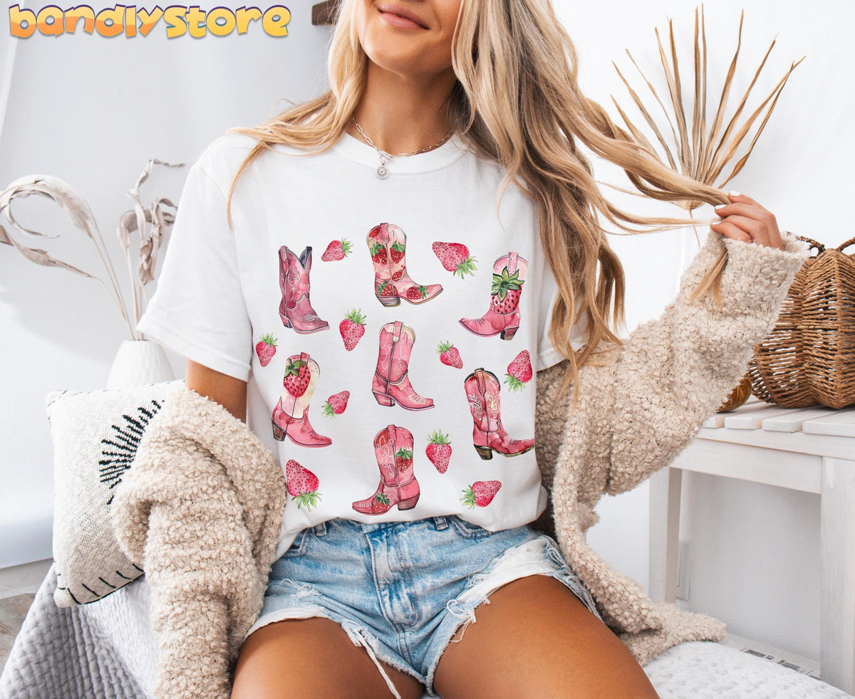 Strawberry Cowgirl Boots T-Shirt on white tee, woman seated indoors amid plants and woven décor.