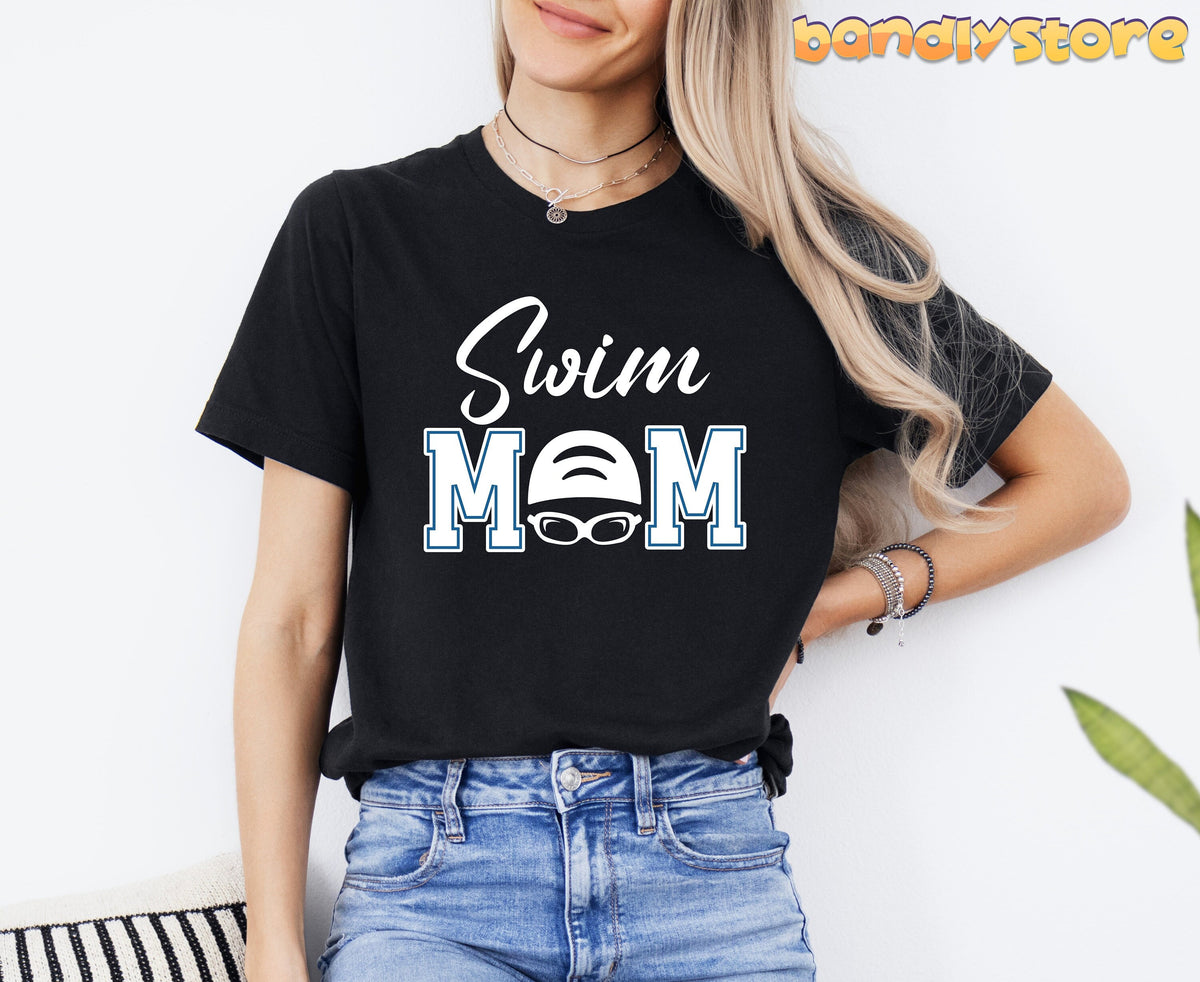 Swim Mom T-Shirt in black on a woman standing against a white wall with jeans, plant and striped cushion nearby.