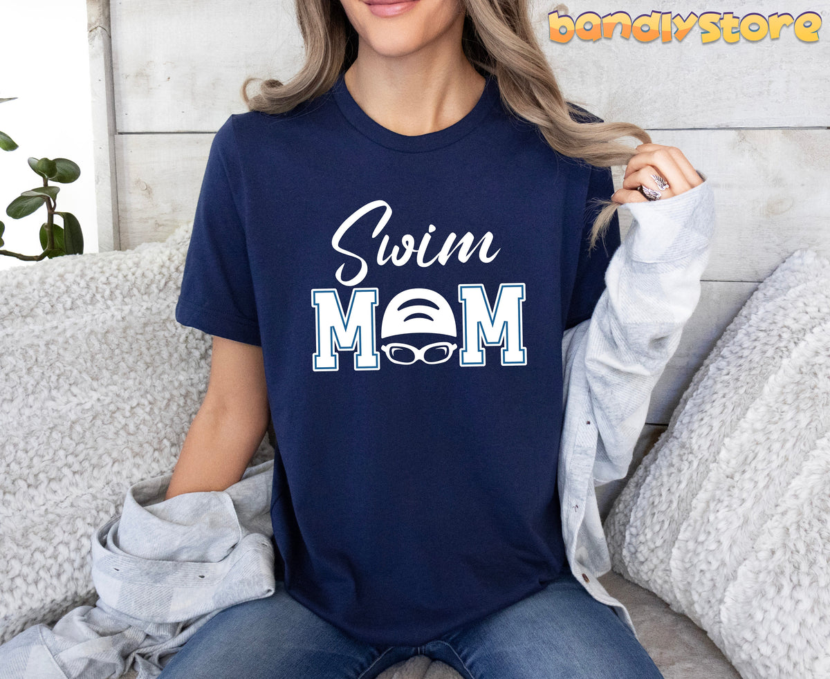 Swim Mom T-Shirt in navy worn by a seated woman on a white couch against wood paneling with a plant.