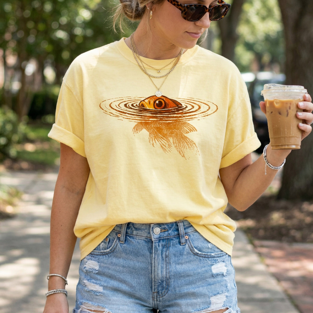 Swimming Goldfish T-Shirt in butter worn by a woman outdoors holding an iced coffee amid trees and a path.