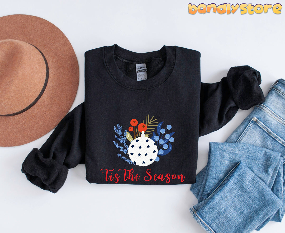 Tis The Season Sweatshirt in black folded flat beside a brown hat and blue jeans.
