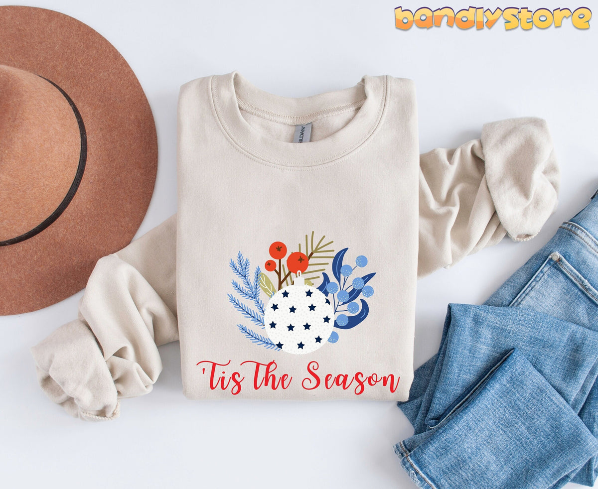 Tis The Season Sweatshirt in sand folded flat next to a brown hat and blue jeans on white.