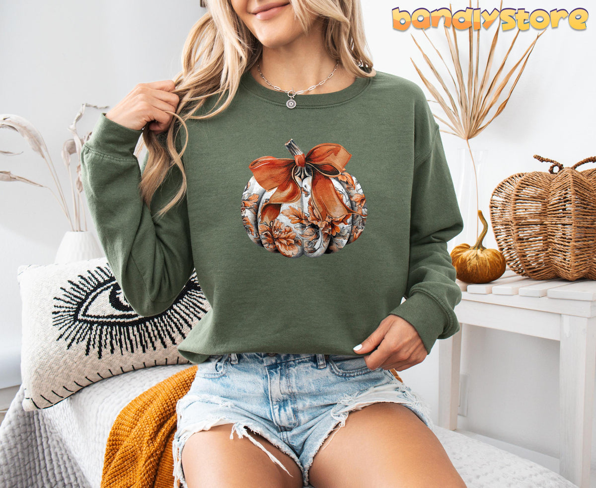 Cute Toile Pumpkin Bow Sweatshirt in military green, modeled front-facing indoors beside dried plants, woven pumpkins and a patterned cushion.