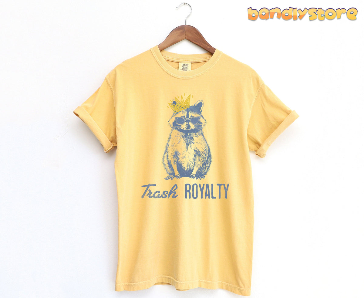 Trash Royalty Raccoon T-Shirt in mustard hangs on a wooden hanger against a plain white wall.