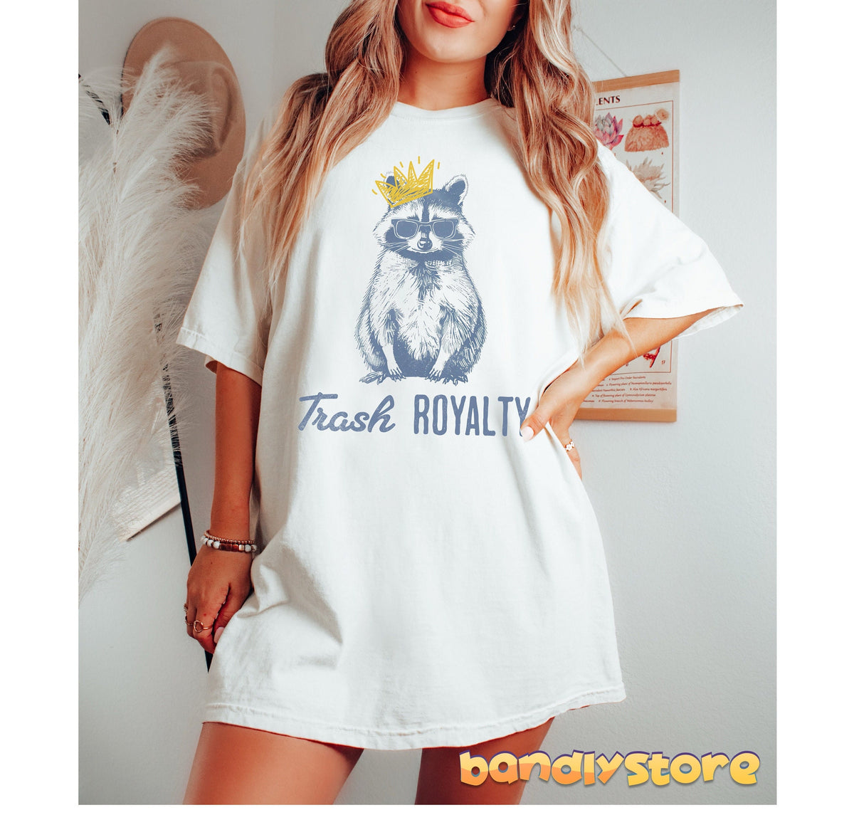 Trash Royalty Raccoon T-Shirt in white on a model standing front-facing beside hat, pampas grass and wall poster.