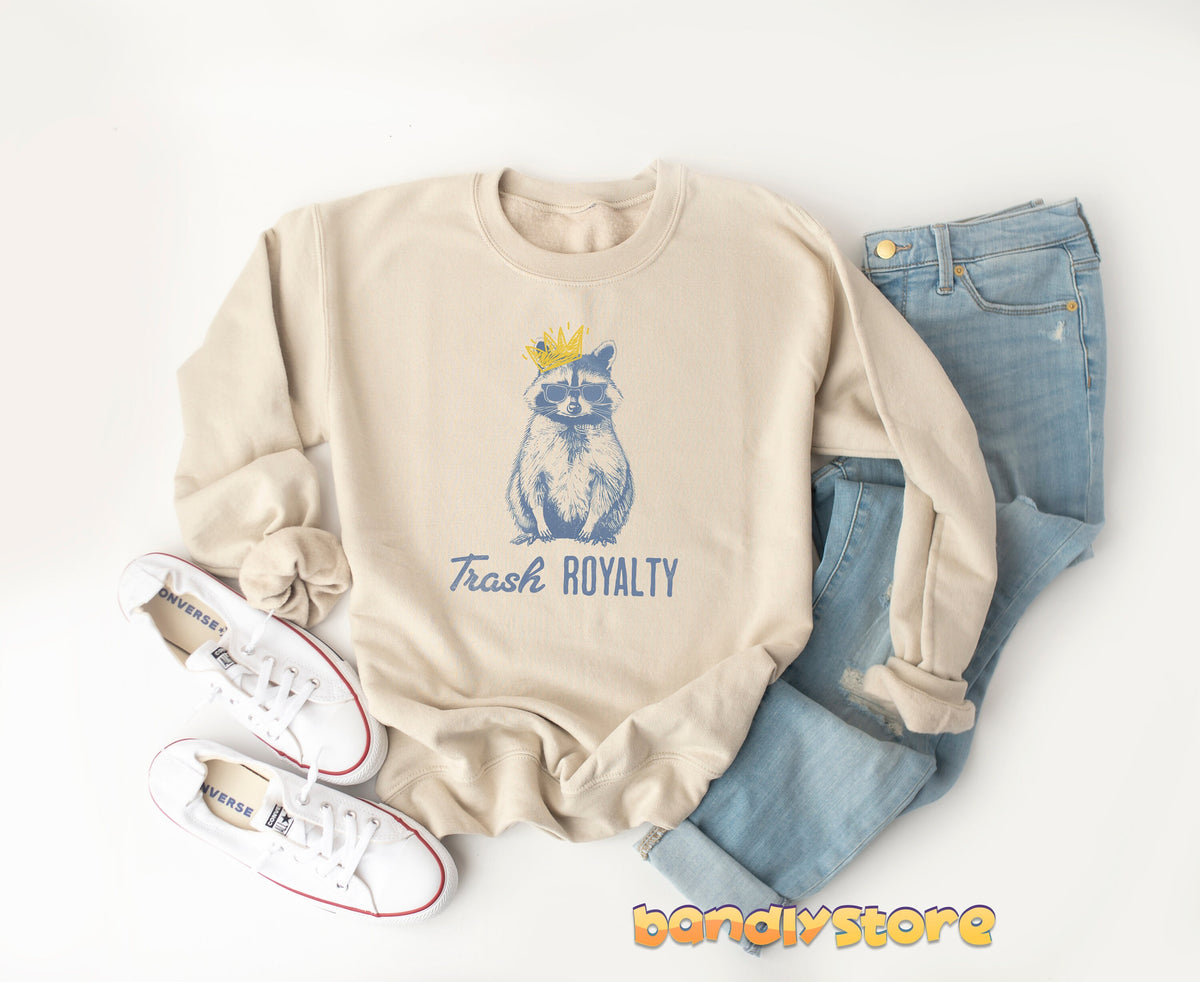 Trash Royalty Raccoon Sweatshirt in sand flat-laid beside white sneakers and blue jeans.