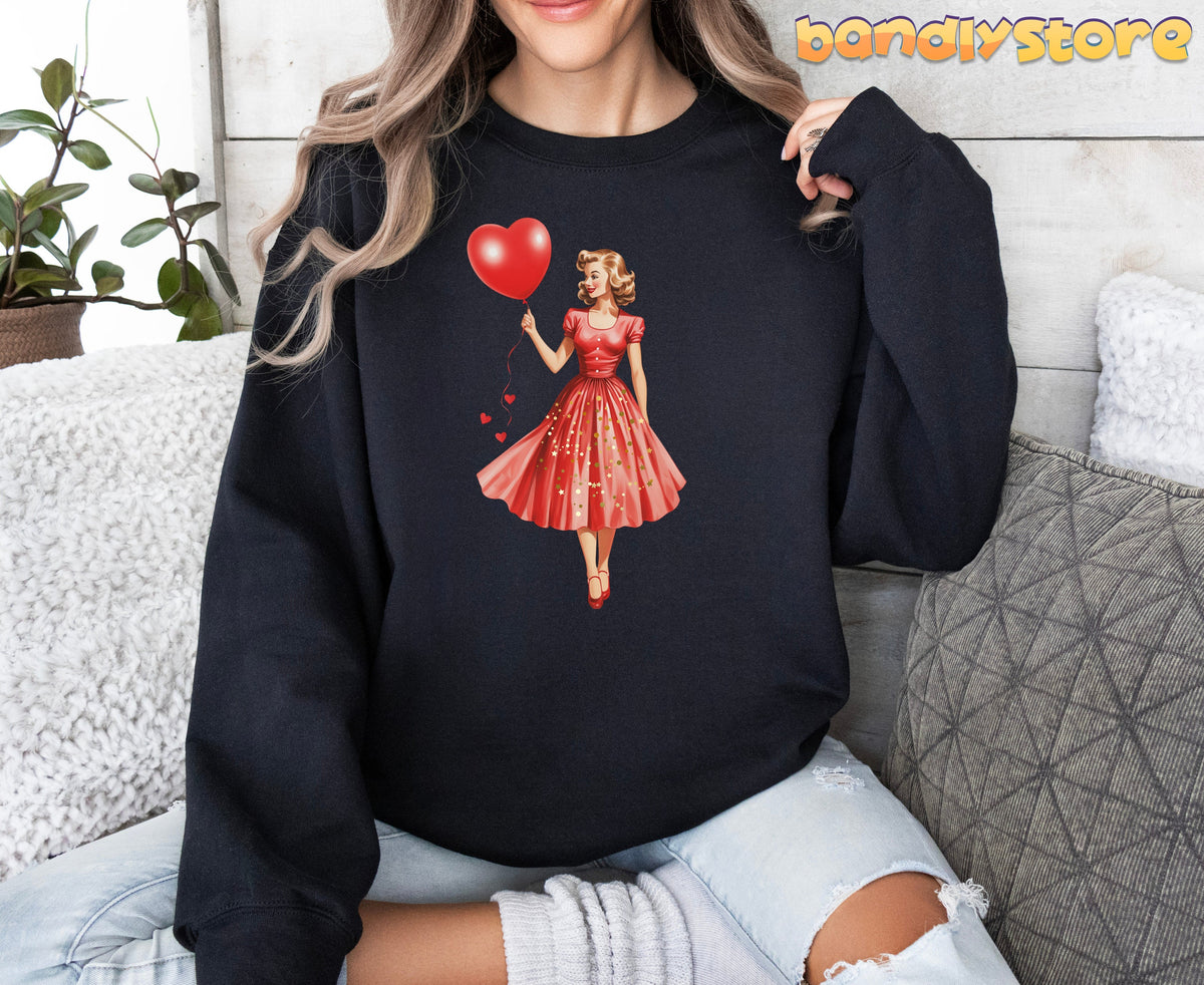 Valentine Girl Sweatshirt in black, worn by a seated woman amid cushions and a plant against a wood wall.