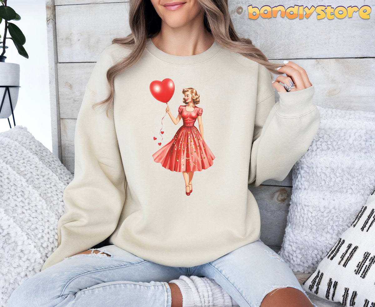 Valentine Girl Sweatshirt in sand worn by a seated model against a wooden wall with pillows and a plant.