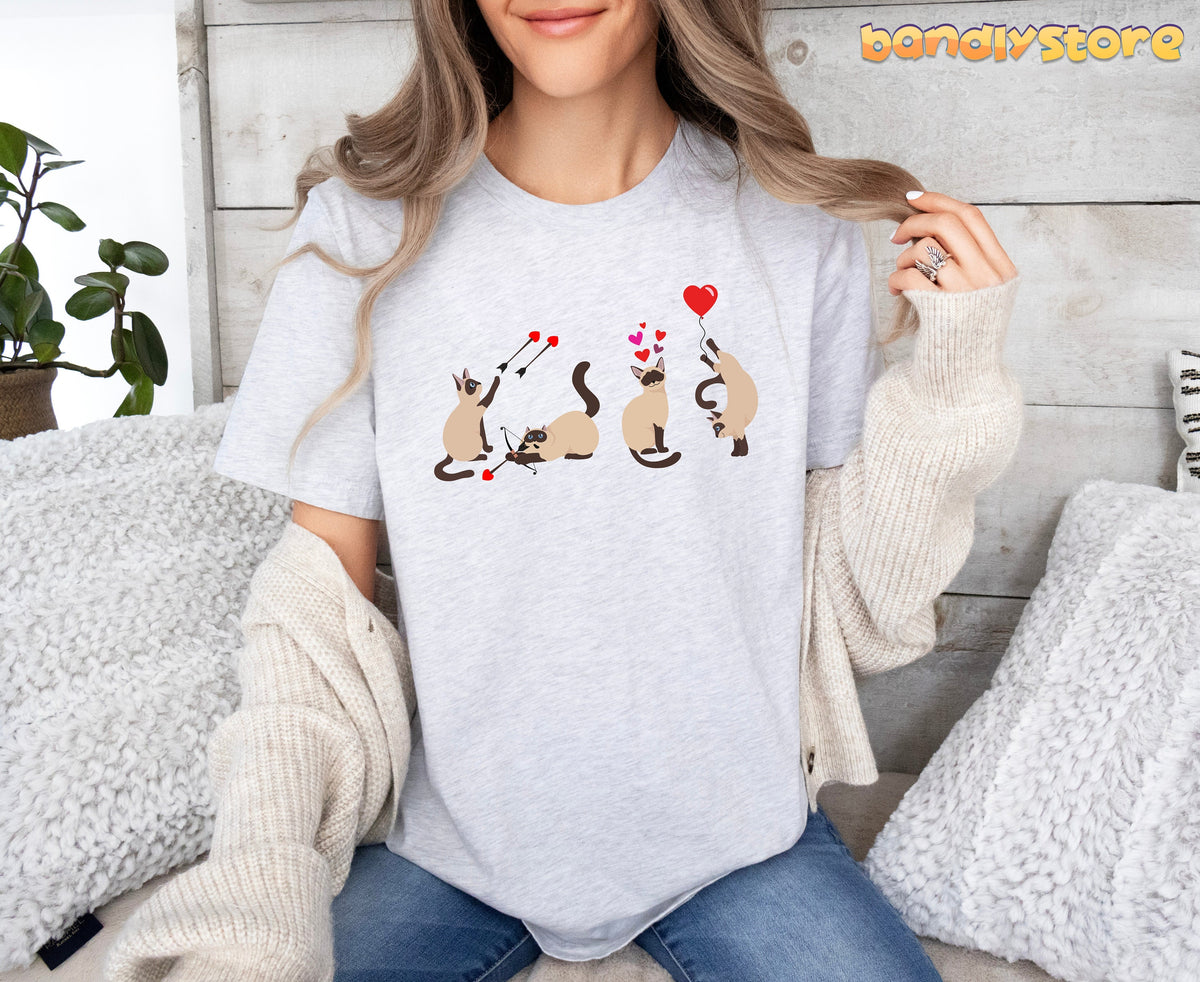 Valentine's Day Siamese Cat T-Shirt in ash, front view on seated model next to plant and pillows against wooden wall.
