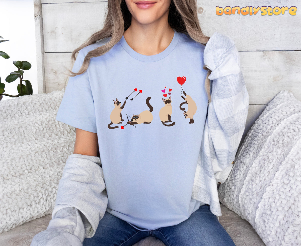 Valentine's Day Siamese Cat T-Shirt in light blue on a person seated on a couch with pillows against a wooden wall.