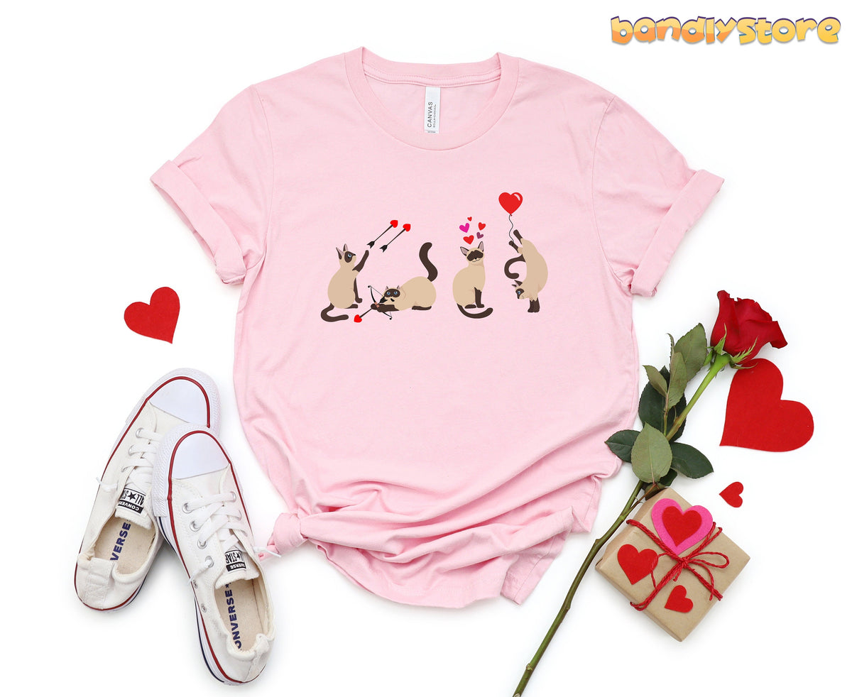Valentine's Day Siamese Cat T-Shirt in pink, flat-lay with white sneakers, red rose, gift box and scattered hearts.