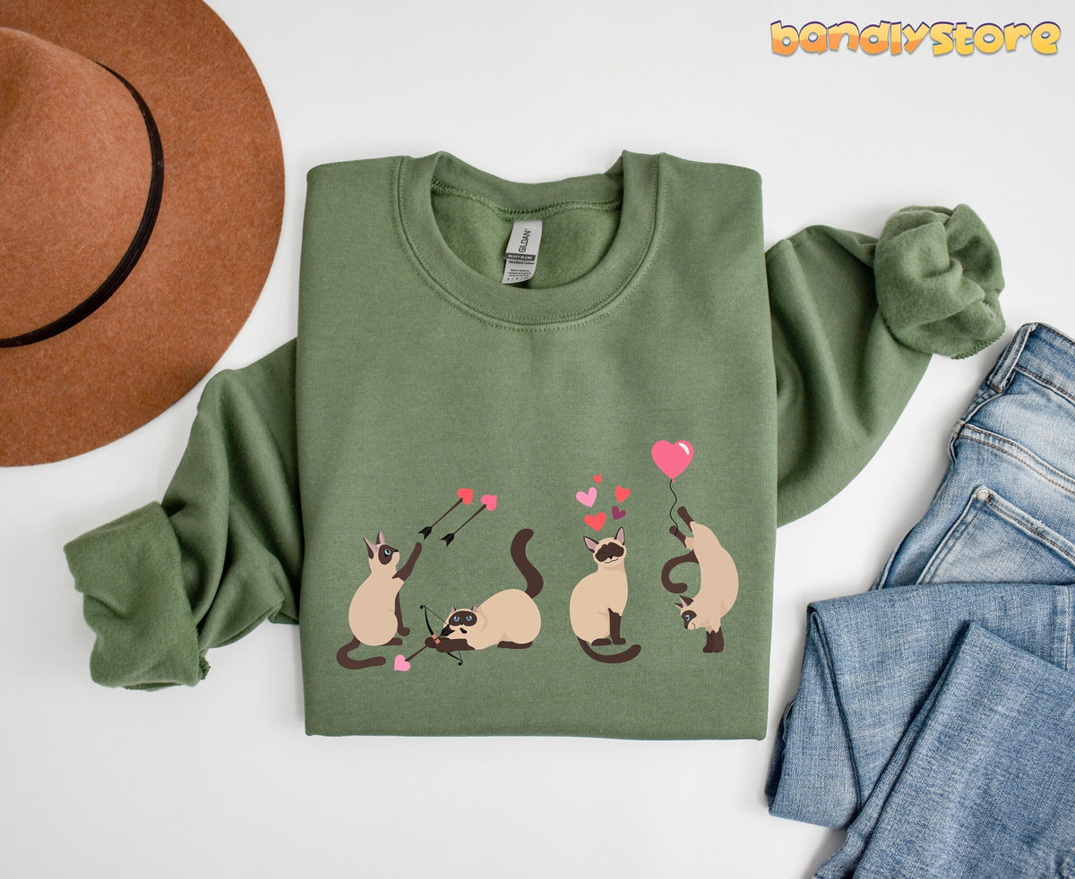Siamese Cat Valentine Sweatshirt in military green laid flat beside a brown hat and blue jeans on white.
