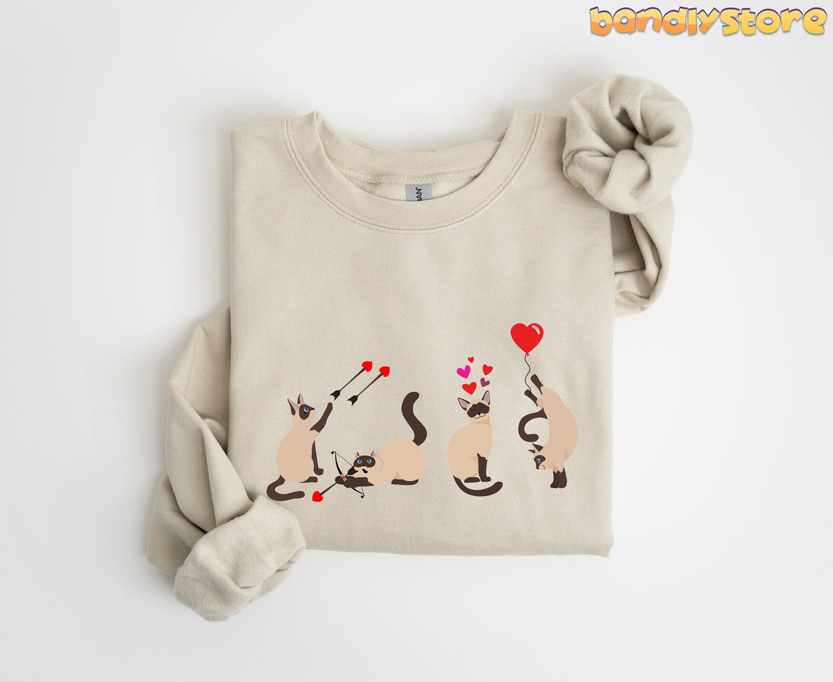 Siamese Cat Valentine Sweatshirt folded on white surface in sand, cats with cupid bows and heart balloon.