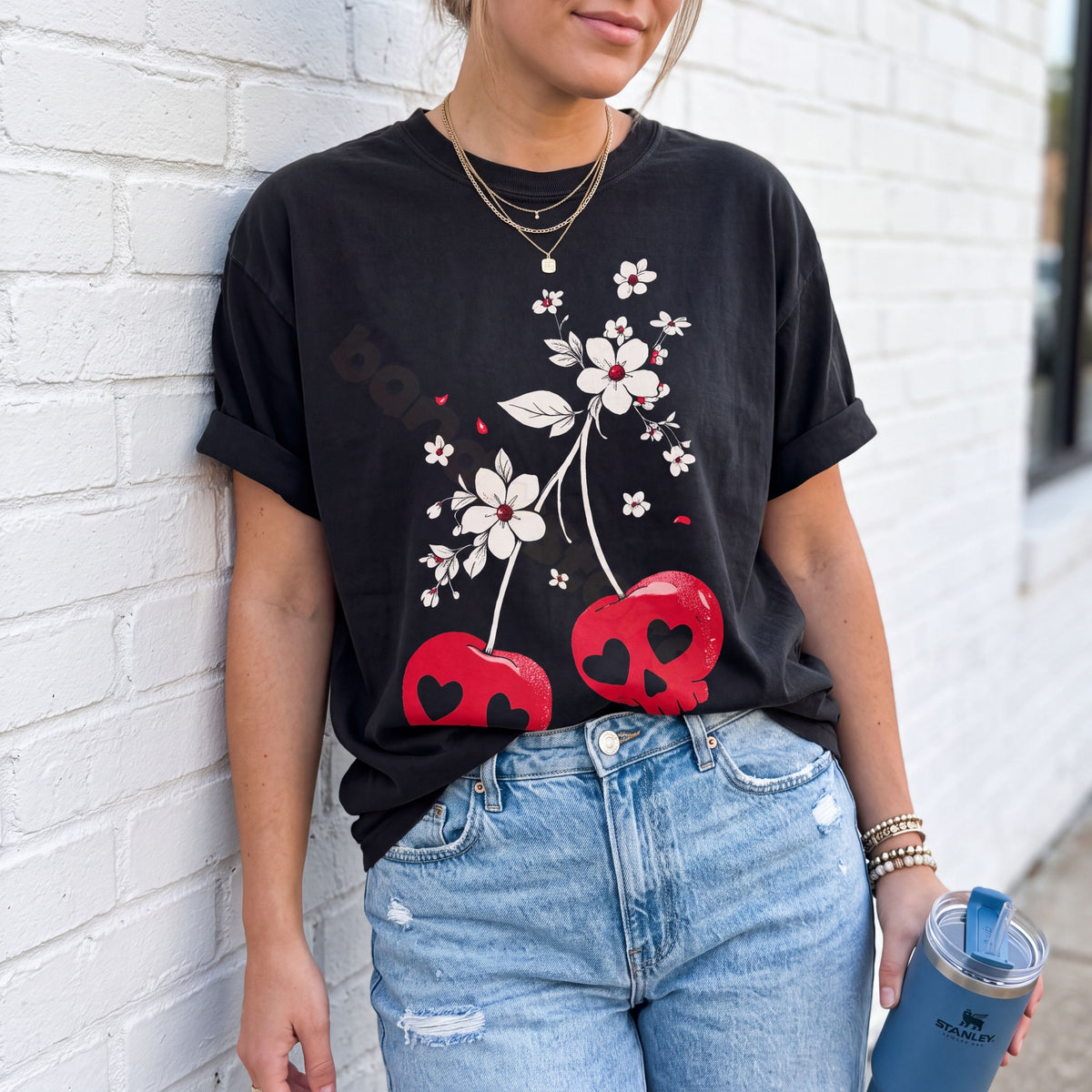 Vintage Cherry Skulls T-Shirt in black, worn front-facing by a person against a white brick wall holding a blue tumbler.