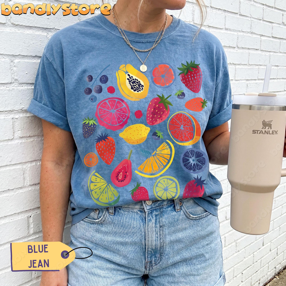 Cute Fruit T-Shirt in Blue Jean on a person against a white brick wall holding a Stanley tumbler.