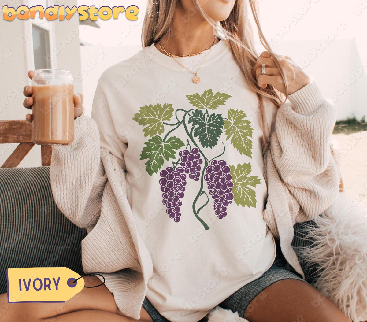 Vintage Grapes T-Shirt in ivory on a woman seated outdoors holding a glass of coffee.
