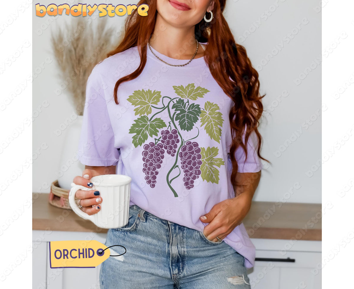 Vintage Grapes T-Shirt in orchid on a woman holding a white mug at a kitchen counter, front view.
