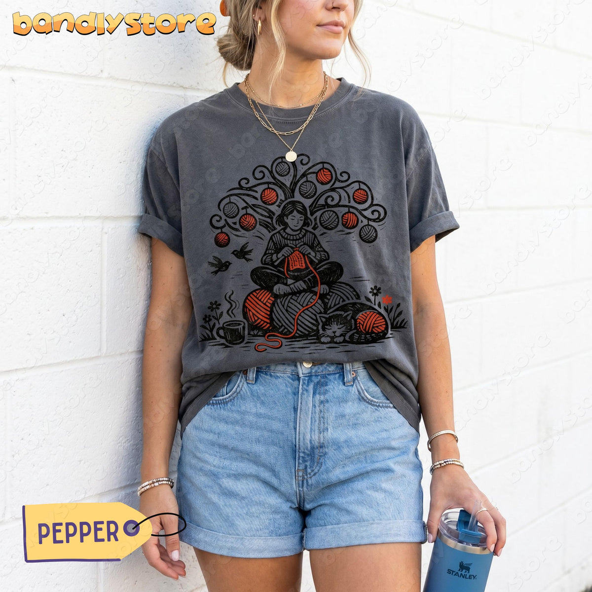 Vintage Linocut Knitting T-Shirt in pepper on a model leaning against a white brick wall holding a tumbler.