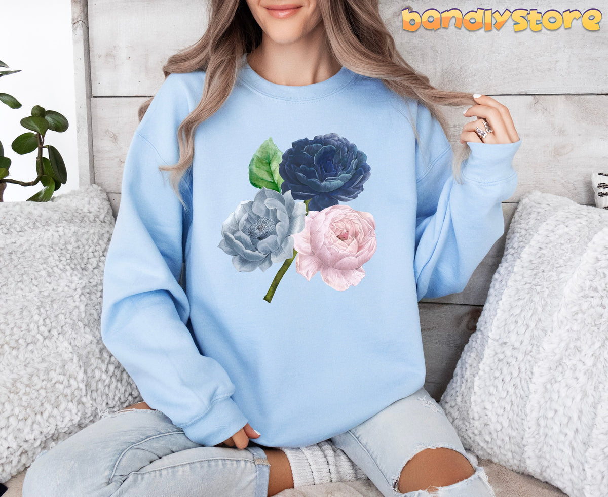 Vintage Roses Sweatshirt in light blue, front view on seated model amid pillows, plant and wooden wall.