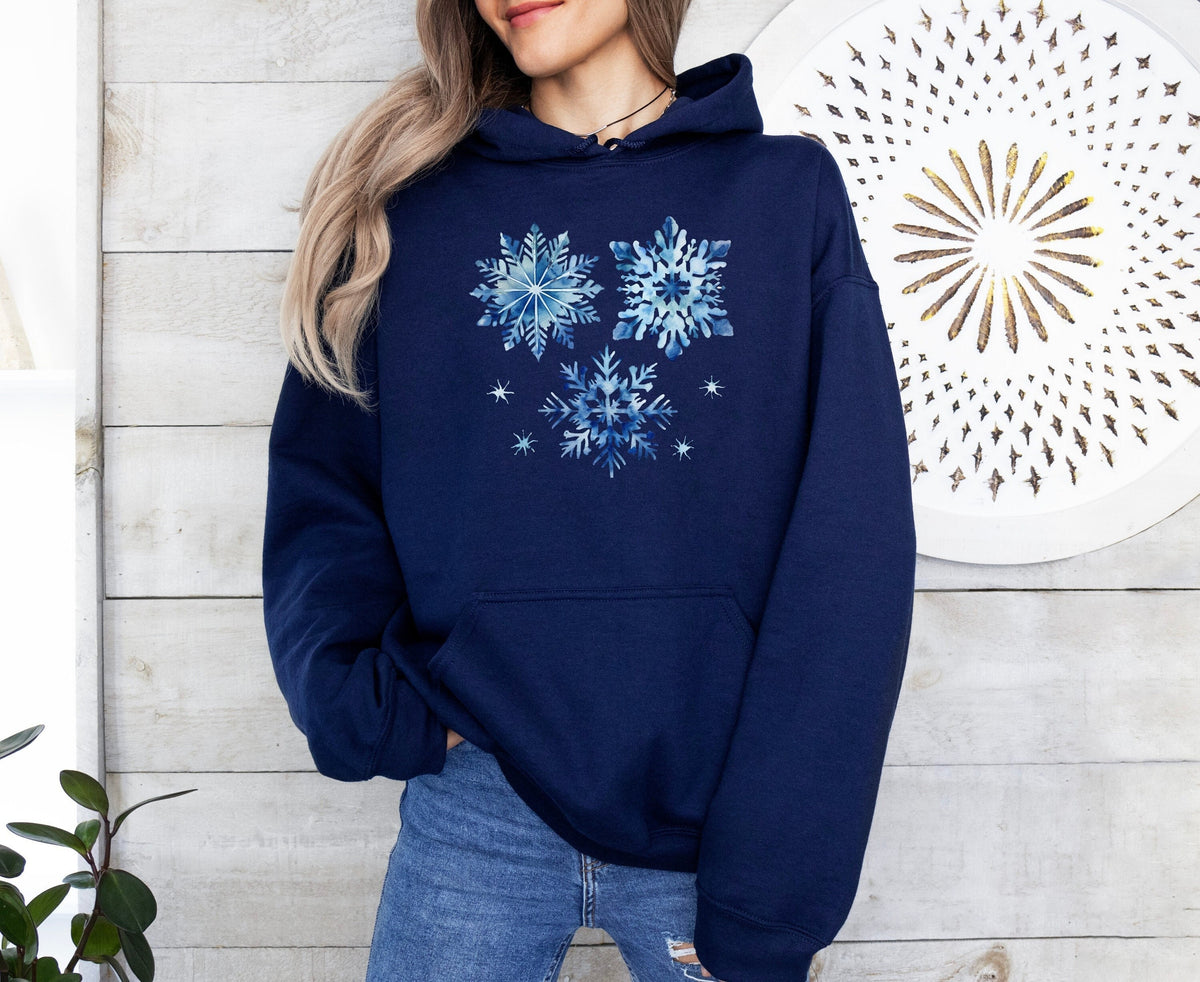 Watercolor Snowflakes Hoodie in navy shown front-on against a light wooden wall with plant and round decorative art.