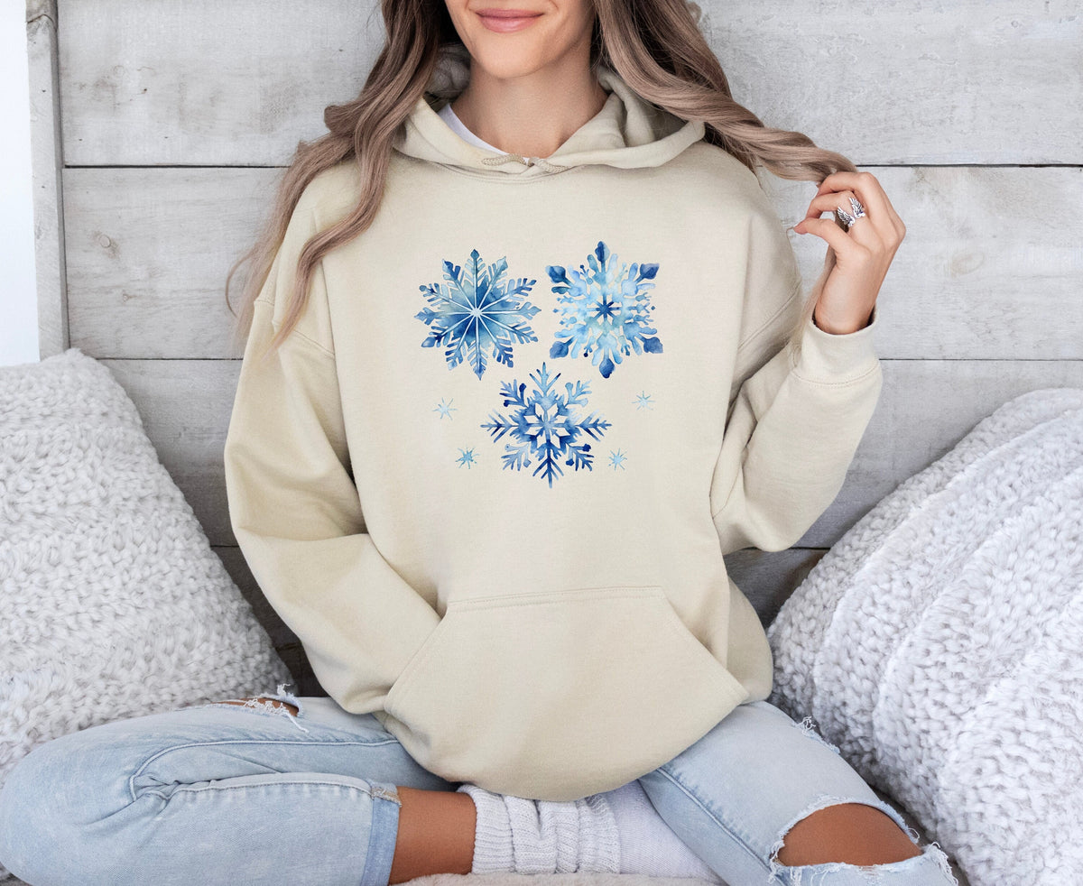 Watercolor Snowflakes Hoodie in sand on a seated person against a wooden wall with white pillows.