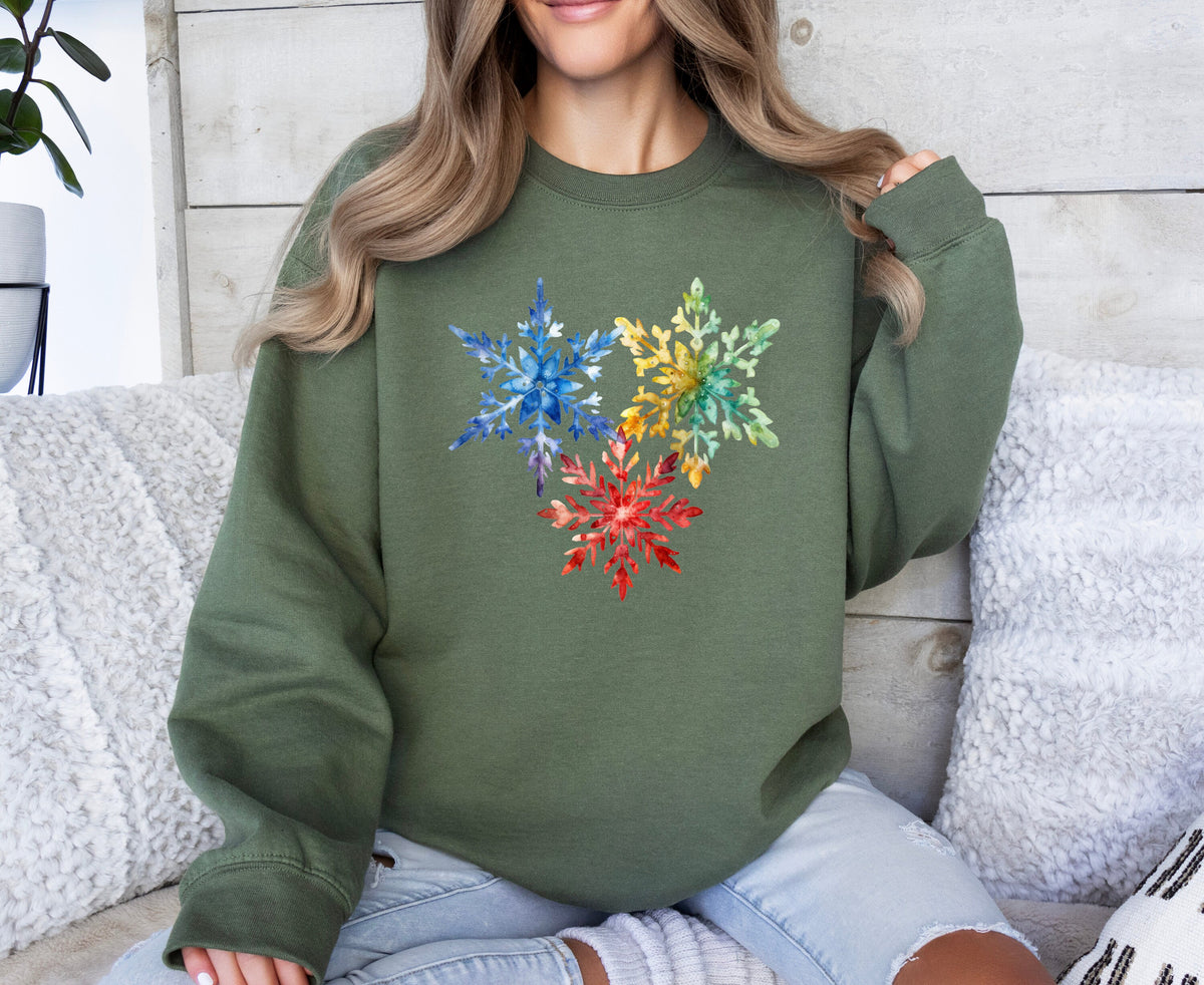 Colorful Watercolor Snowflakes Sweatshirt in military green, worn front-facing on a seated person amid white cushions and a plant.