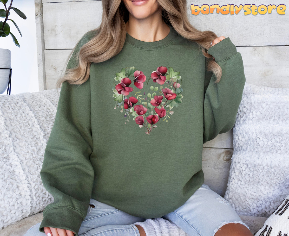 Watercolor Sweet Pea Heart Sweatshirt in military green on a woman sitting on a white-pillowed couch against a wooden wall.
