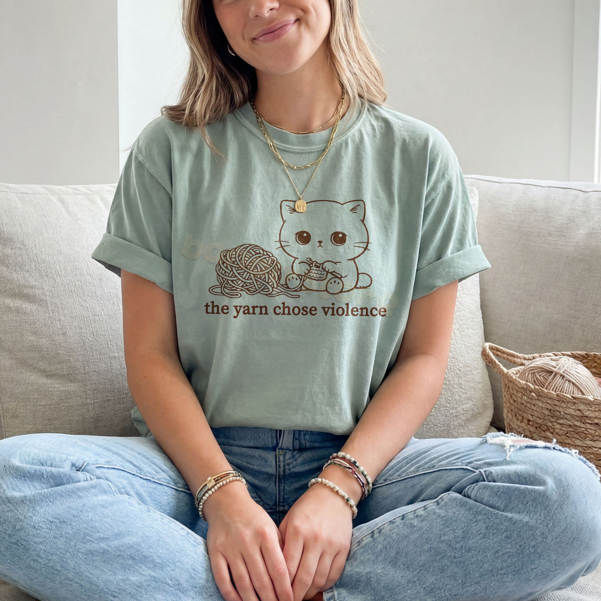 The Yarn Chose Violence T-Shirt in Bay, worn by a person seated cross-legged on a sofa beside a yarn basket.